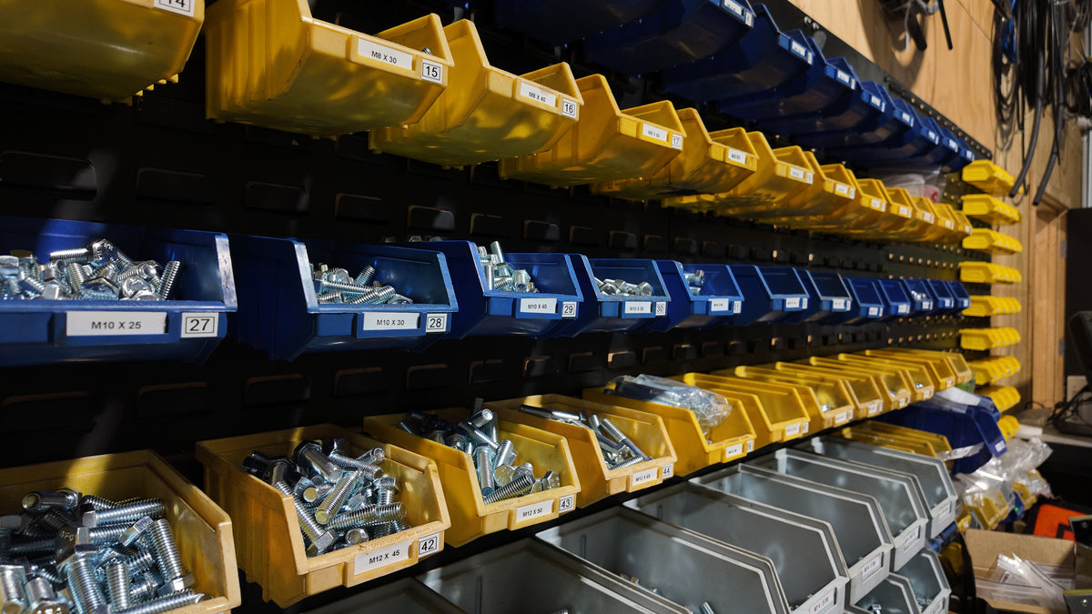 Industrial wall-mounted storage shelves filled with yellow and blue plastic parts bins containing screws, bolts, and nuts, with metal drawers below.