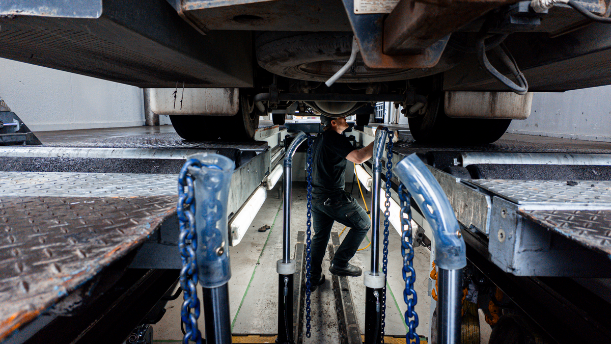 A mechanic in dark clothing walks under a vehicle on a lift, passing blue chains and metal guide rails in a garage setting.