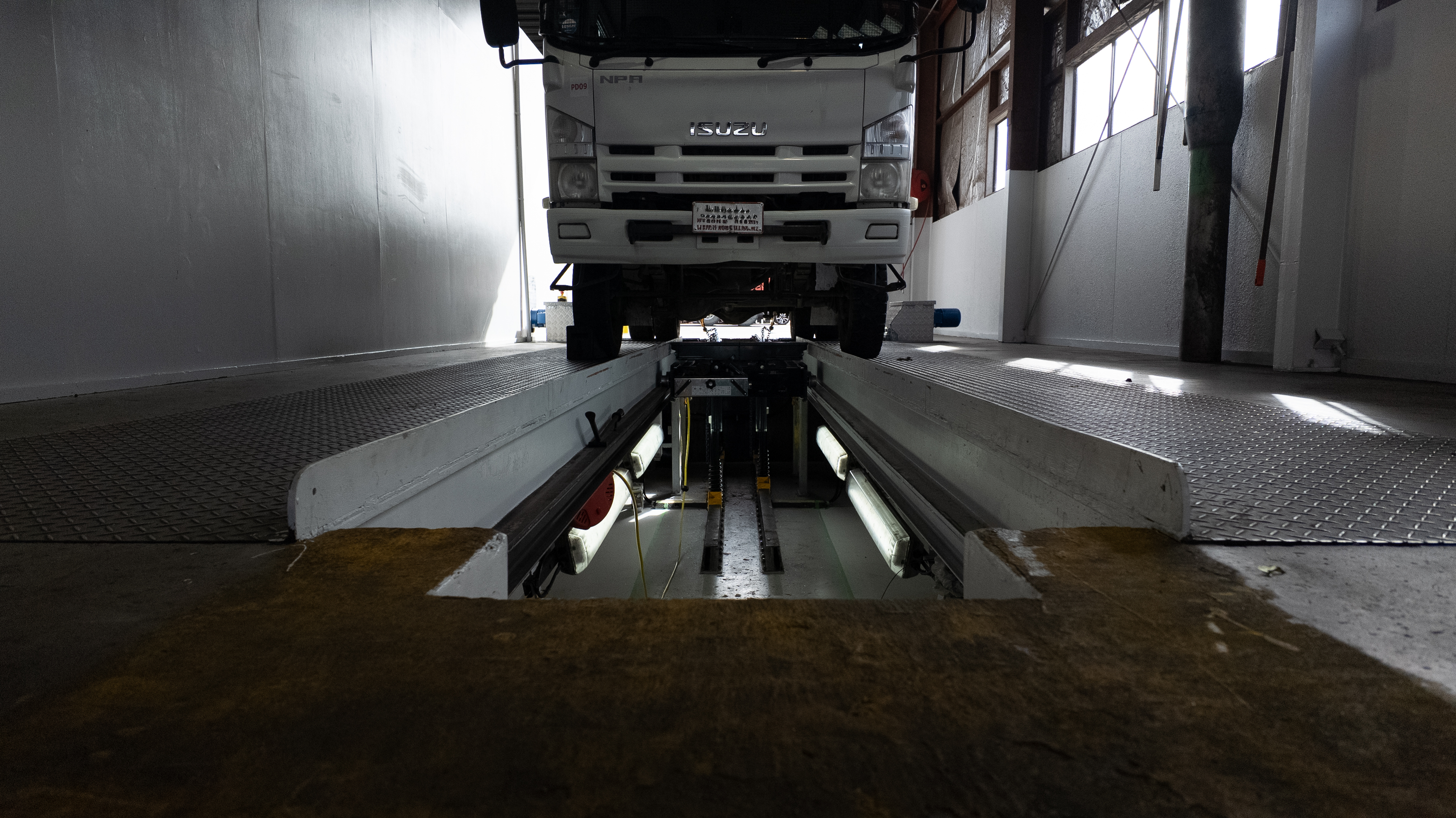 A white Isuzu truck is positioned on a vehicle scale inside a warehouse or testing facility, with metal ramps and industrial walls visible.