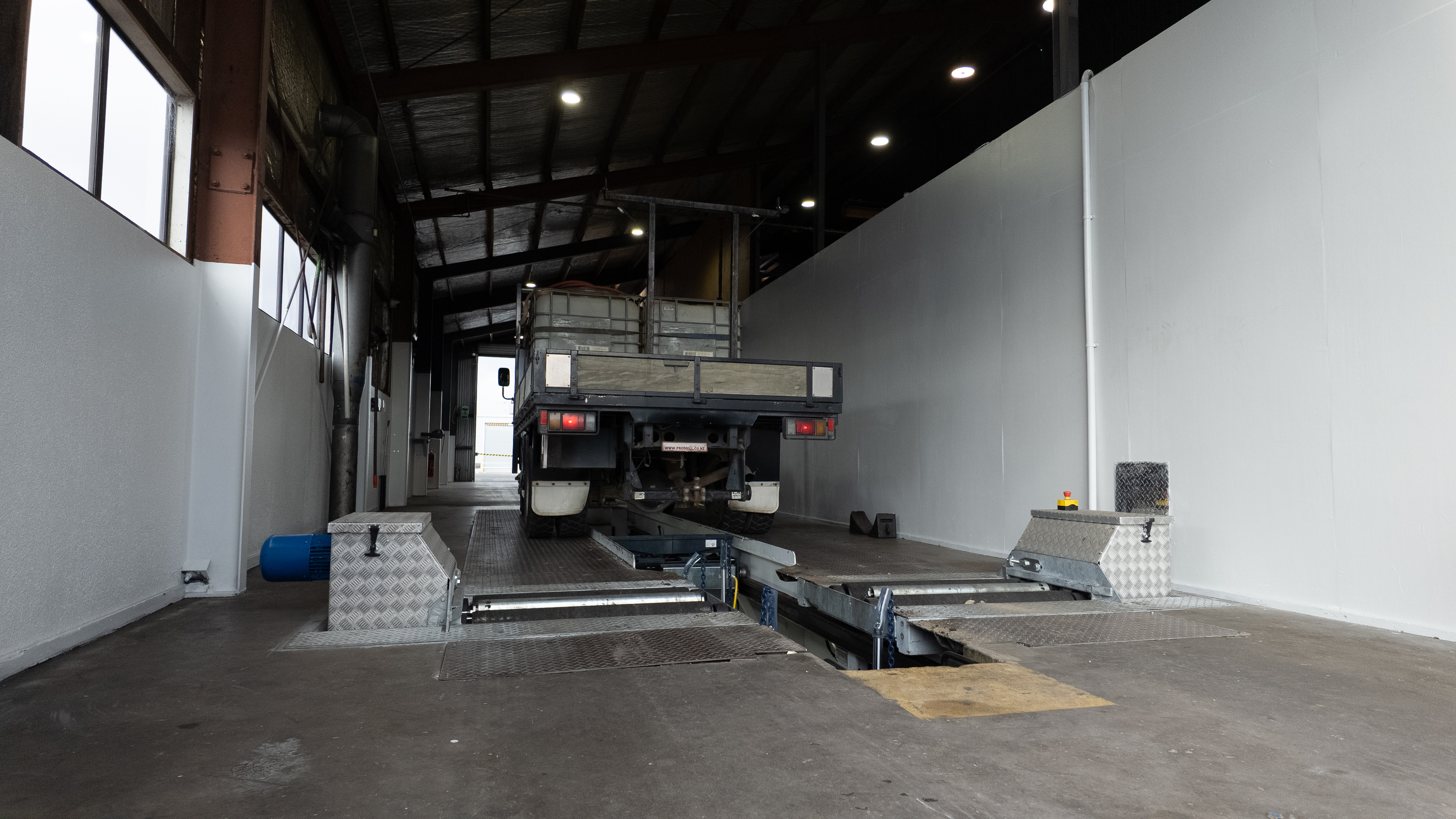 A truck is positioned on a large industrial vehicle lift or gantry inside a warehouse or garage with high ceilings, white walls, and overhead lighting.