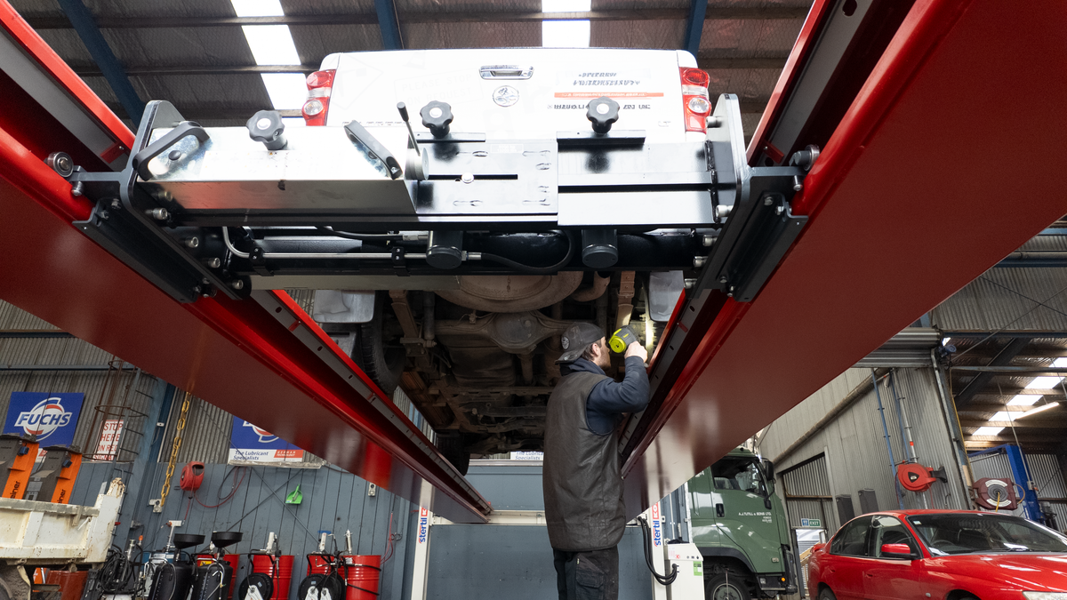 A diesel mechanic in a garage stands underneath a white ute raised on a large red hydraulic lift hoist, inspecting the undercarriage.