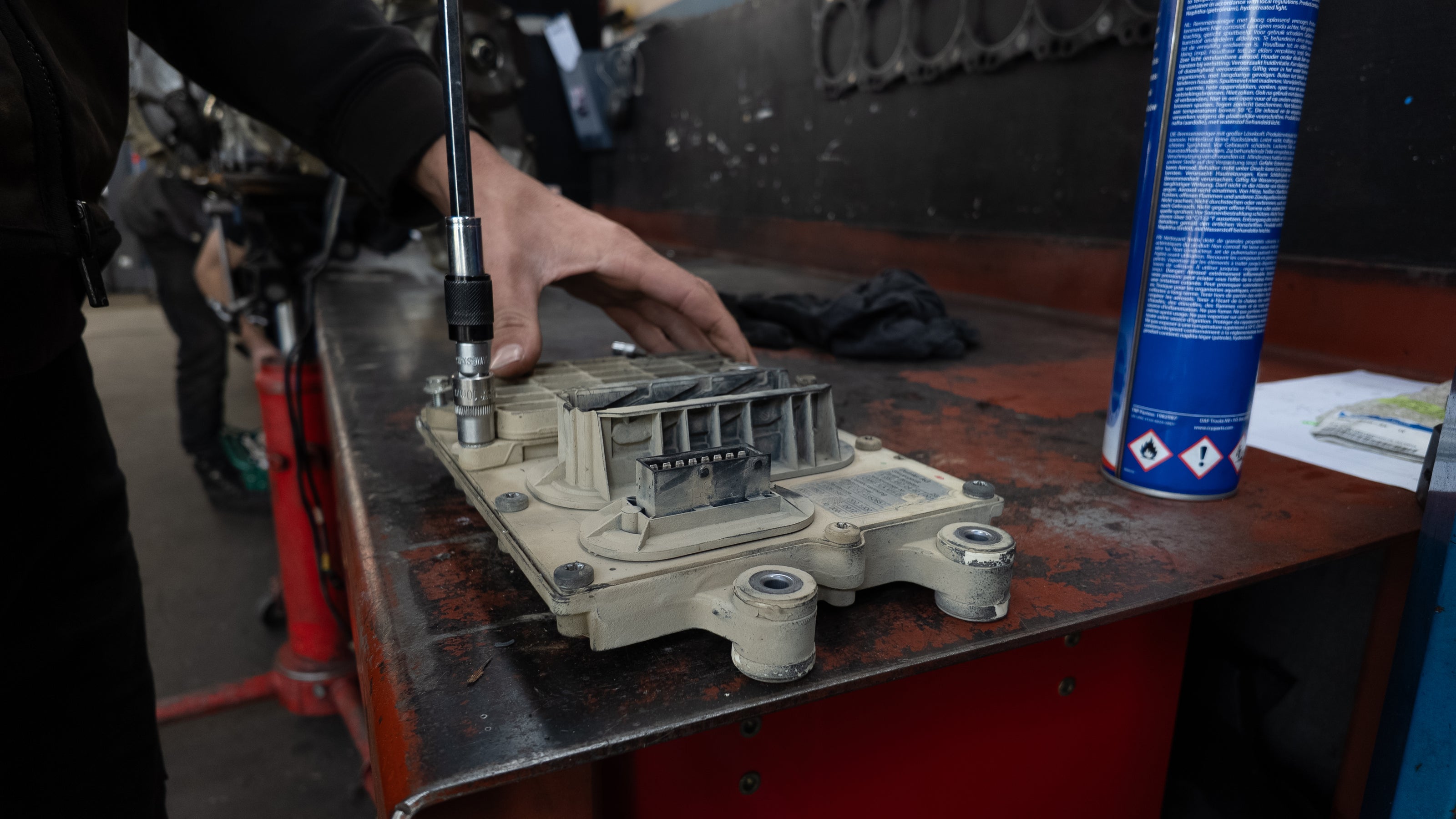 A person's hand using a socket wrench to tighten a metal bolt on a beige ECU Unit  resting on a workbench, with a blue spray can nearby.