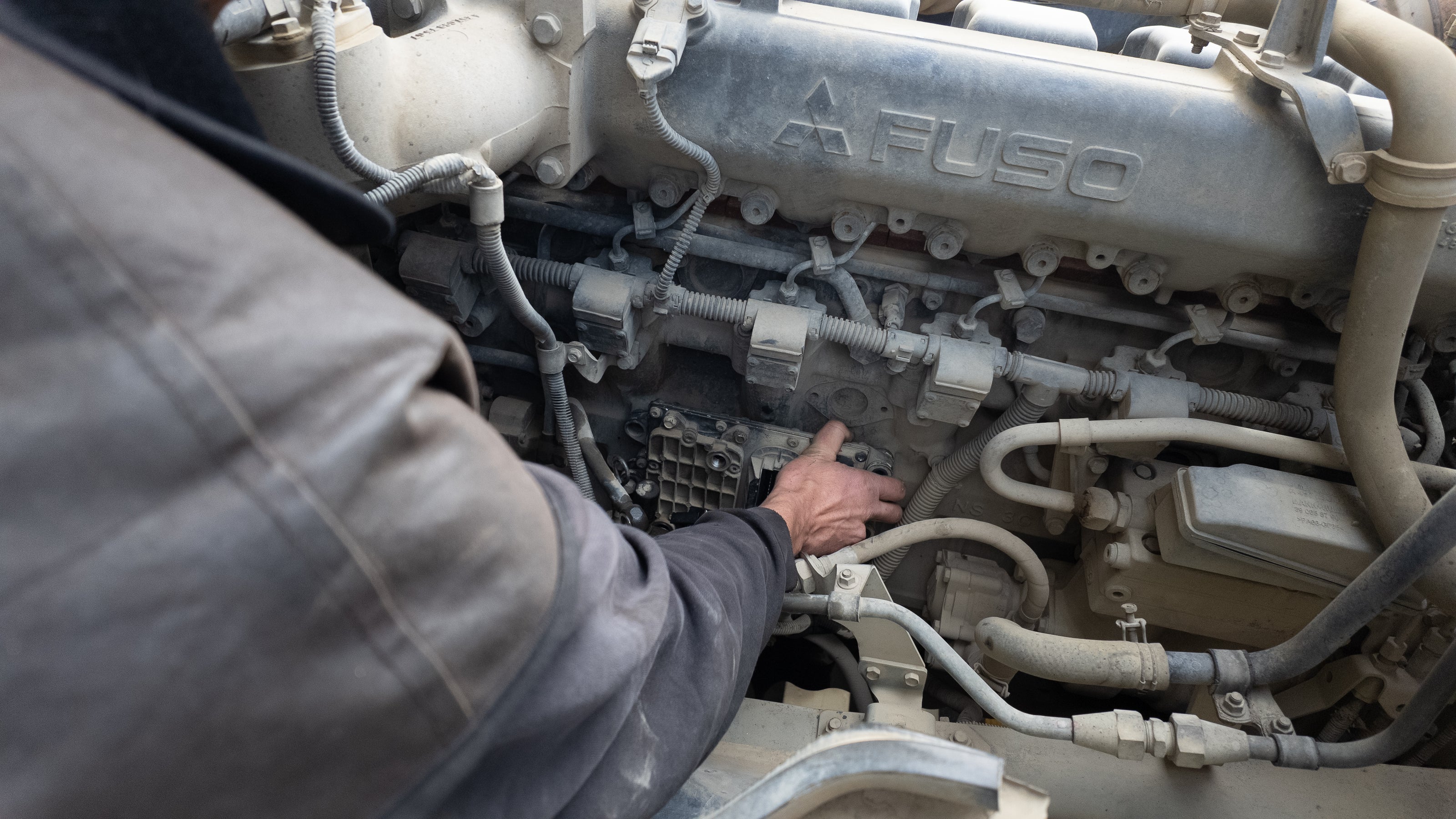 A close-up view of a person's hand pointing to the engine block of a Fuso commercial vehicle, with the silver engine cover and various hoses and components visible including the ECU Unit.