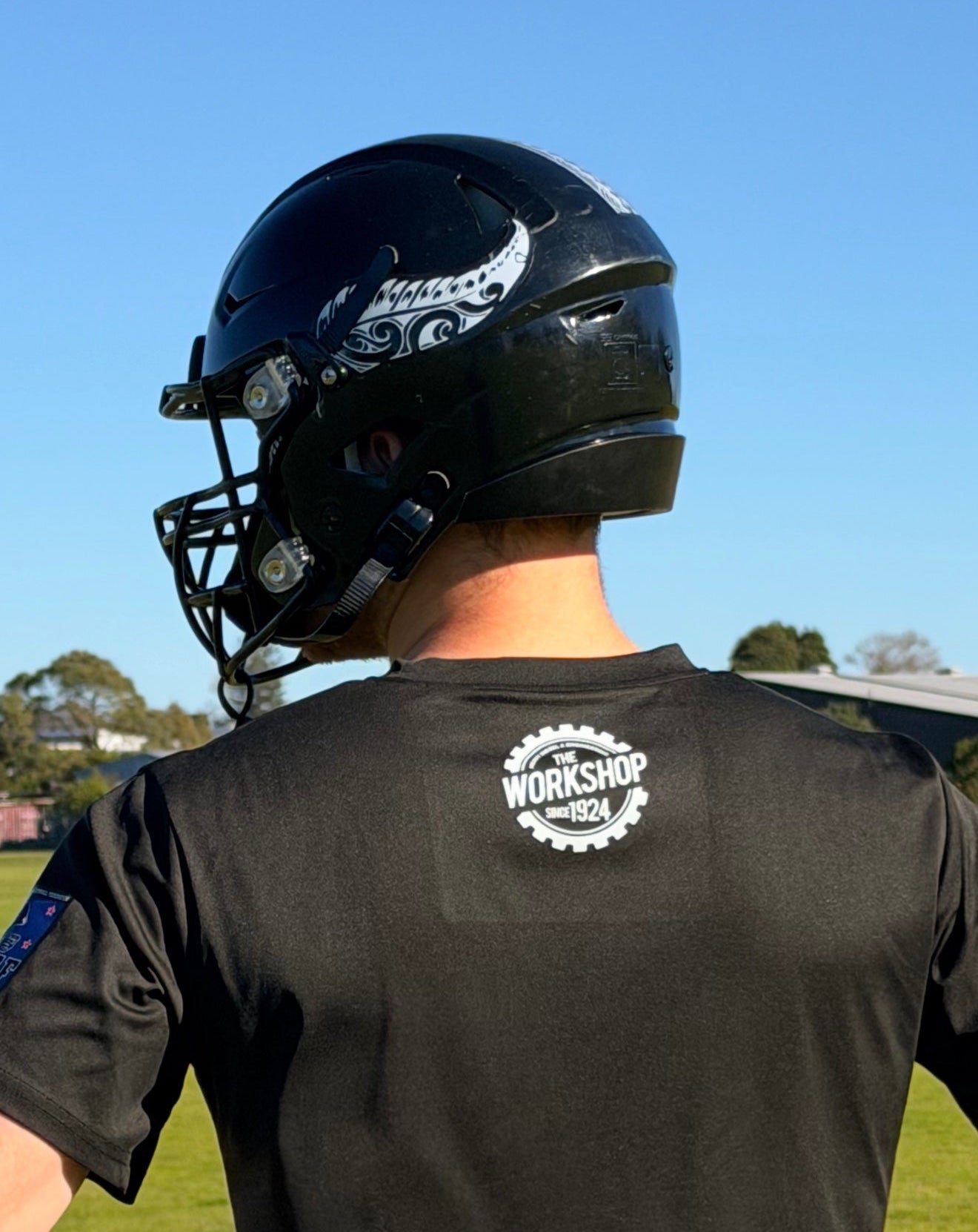 Gabriel Thackham stands outdoors on a grassy field wearing a black football helmet with a face guard and a black T-shirt, photographed from behind. The shirt has a white circular 'The Workshop 1924' logo on the upper back.