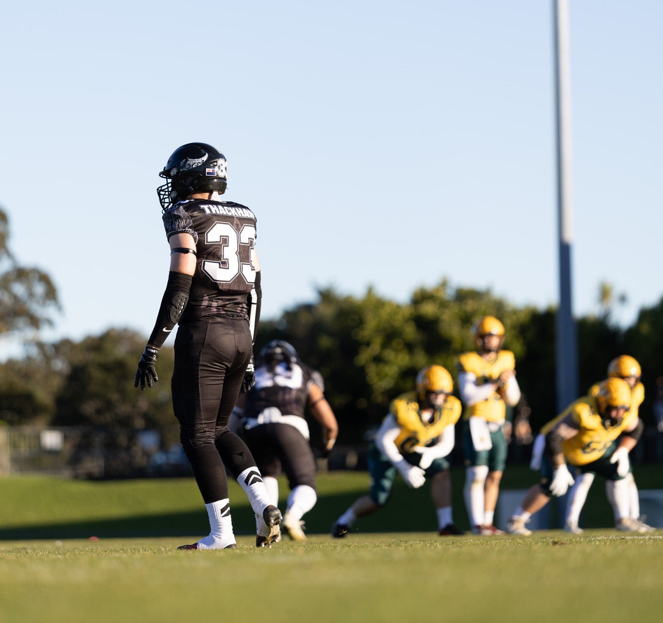 Gabriel Thackham in a black and white uniform with the number 33 stands on a grassy field, with other players in yellow and white uniforms in the background under a tall light pole and blue sky.
