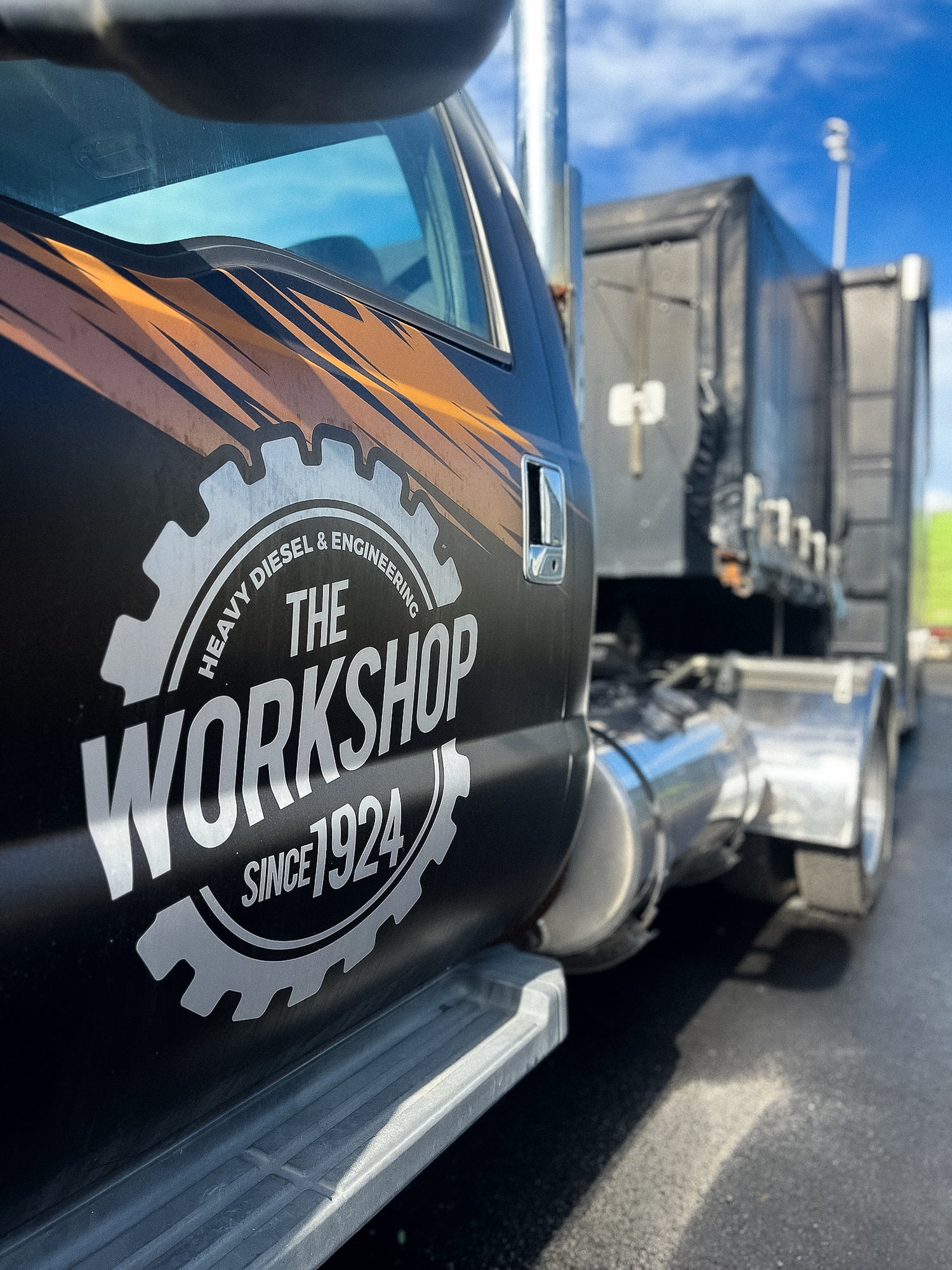 Close-up of a black semi-truck cab with a large white circular 'The Workshop Since 1924' graphic on the side door, parked outdoors under a blue sky.