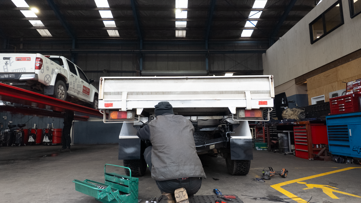 A diesel mechanic in a gray jacket kneels under the rear of a white utility ute inside a large, well-lit diesel workshop. Another white pickup truck is elevated on a red hydraulic lift to the left, and various toolboxes and equipment are visible in the background.