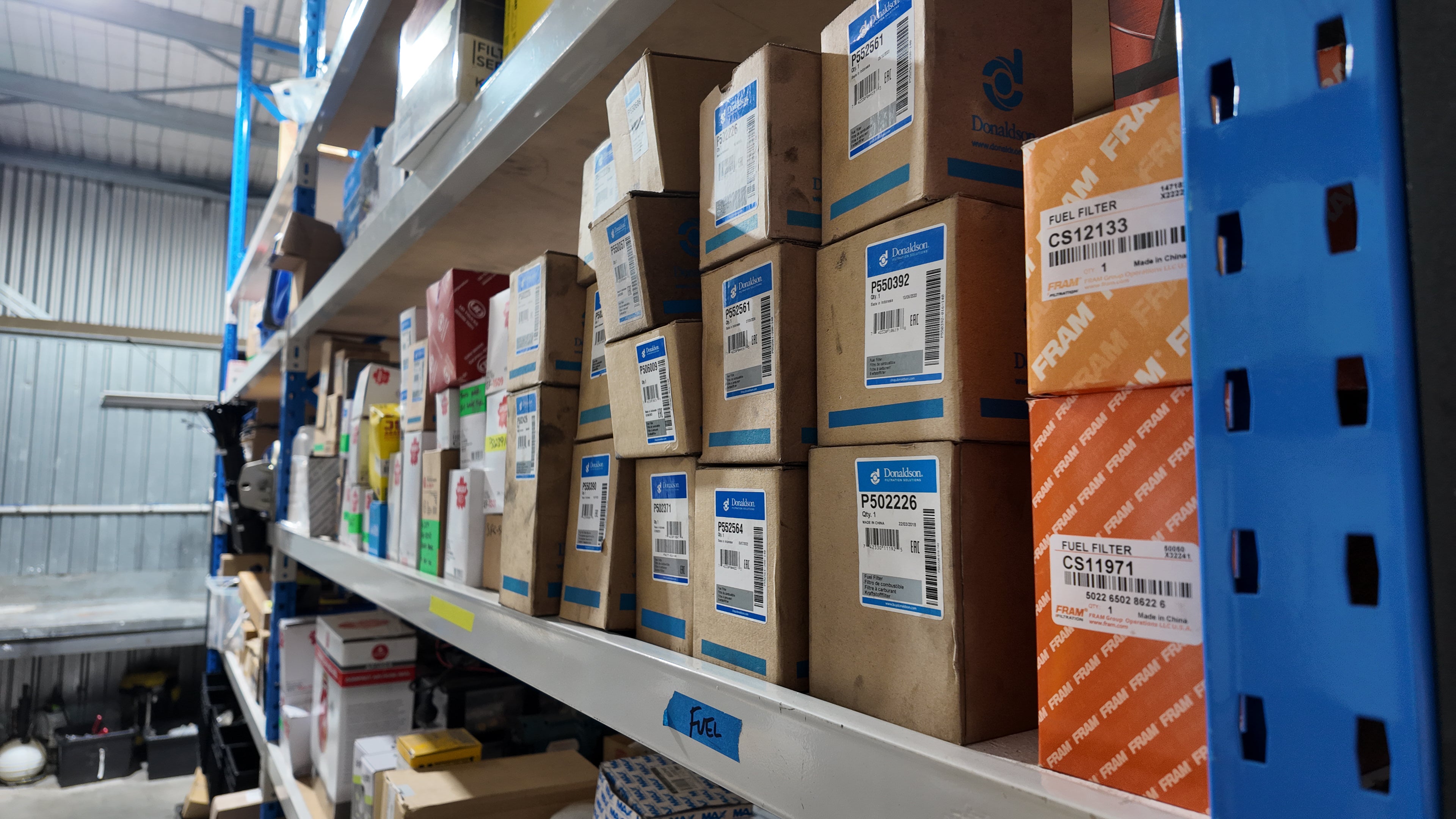 A warehouse aisle with metal shelving stocked with many cardboard boxes. Several boxes show blue and orange branding and printed labels, including visible text such as 'TRAIL' and 'FZG'.