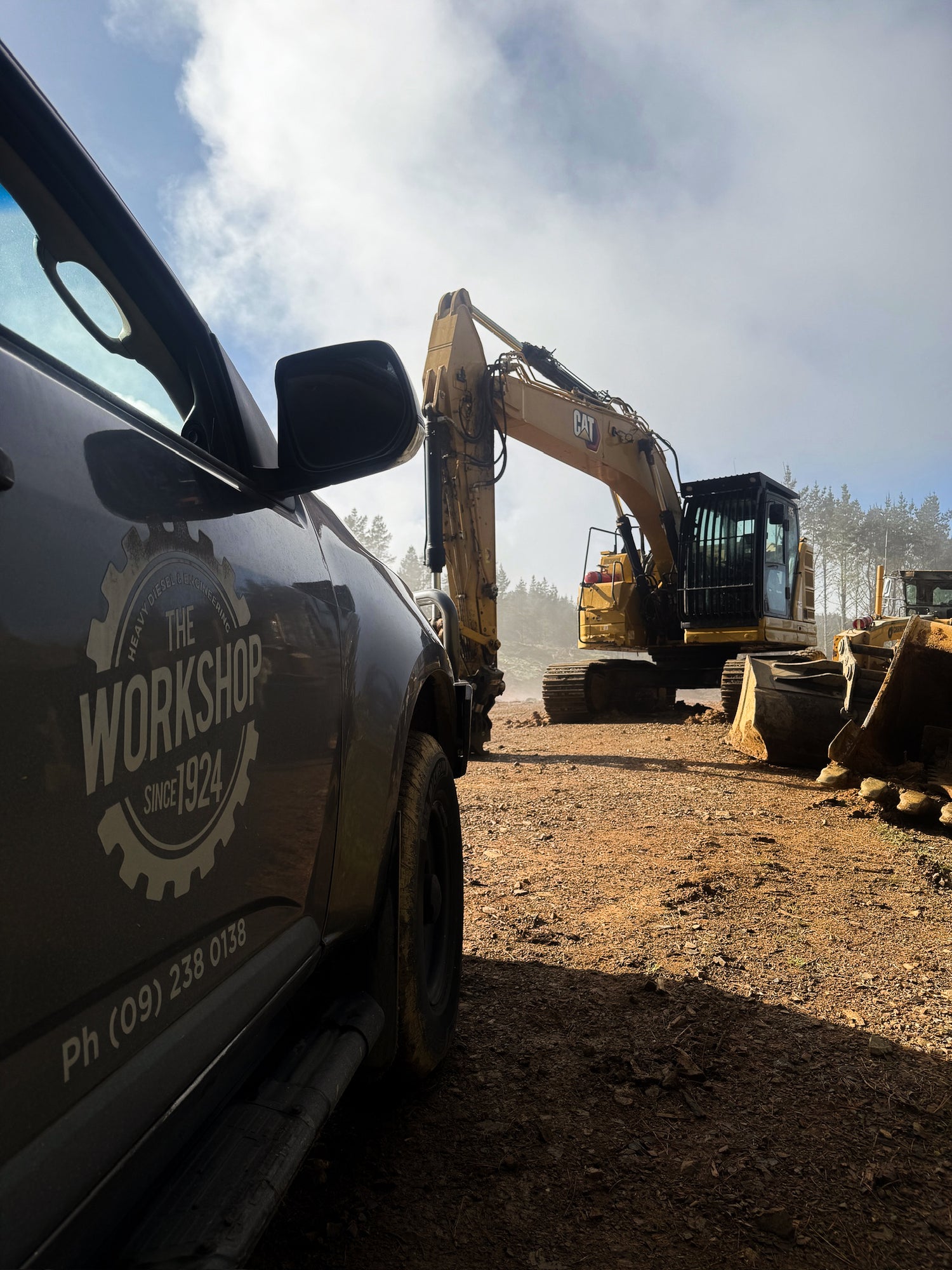 A dark gray vehicle in the foreground shows a large circular logo reading 'THE WORKSHOP SINCE 1924' on its side door, with a dirt construction site and a yellow excavator in the background under a cloudy sky.