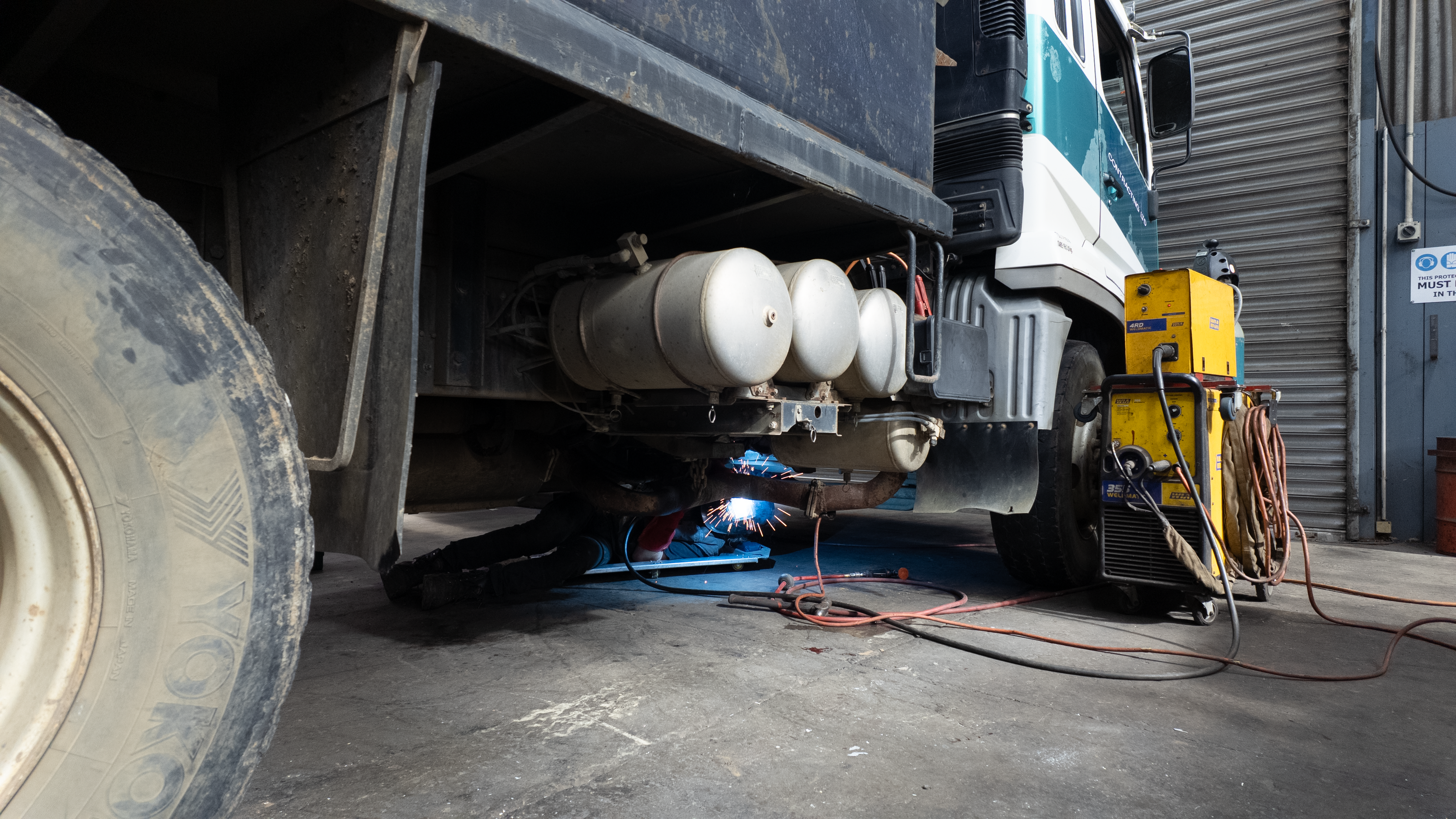A close-up view under a truck showing a welding setup in use, with a bright blue welding arc, white cylindrical tanks, a yellow welding machine, and hoses on a concrete floor in a workshop.
