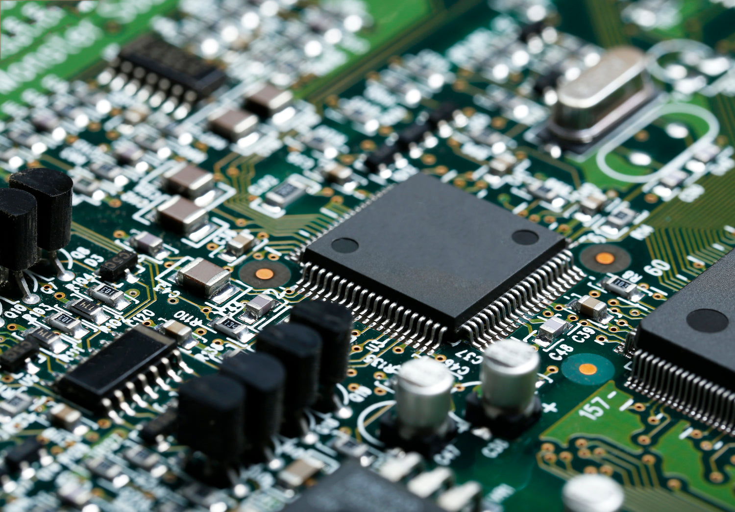 Close-up macro shot of a green printed circuit board (PCB) featuring a central black integrated circuit chip, other electronic components, and fine gold and silver traces.