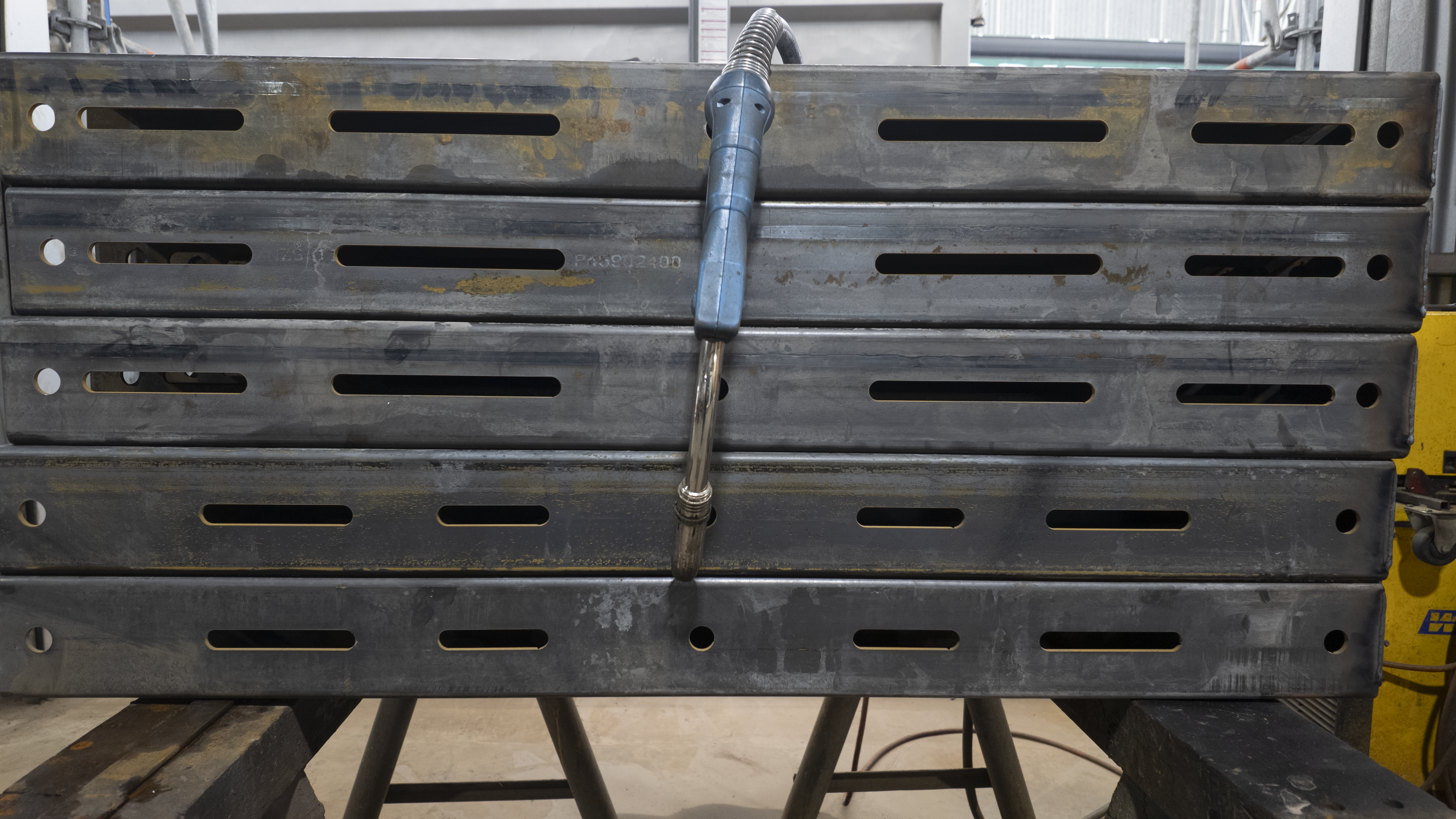 A stack of large, pre-drilled steel plates or deck boards in a workshop setting, with a blue and silver hot air weld gun resting vertically on top.