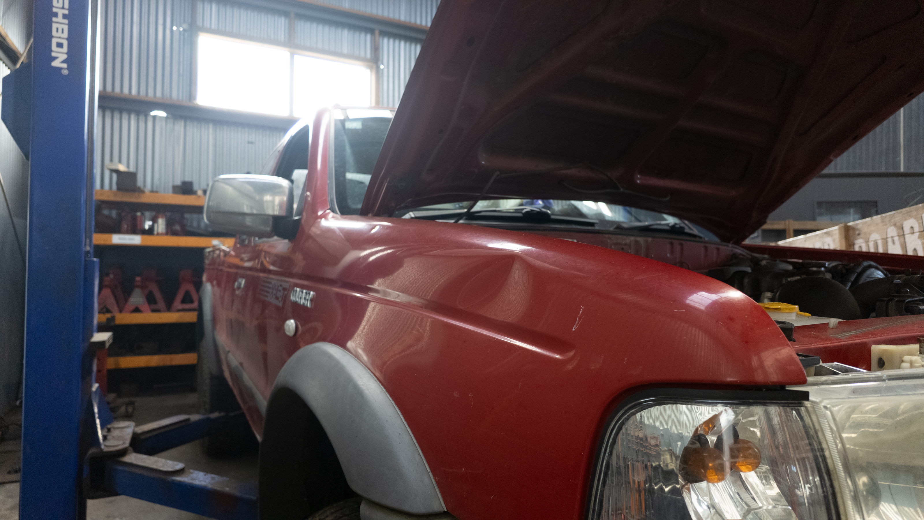 A red Ford courier ute with its hood open, parked inside a garage on a blue vehicle lift. The truck has a visible dent on the front fender and shows a 'COURIER' badge on the side door.