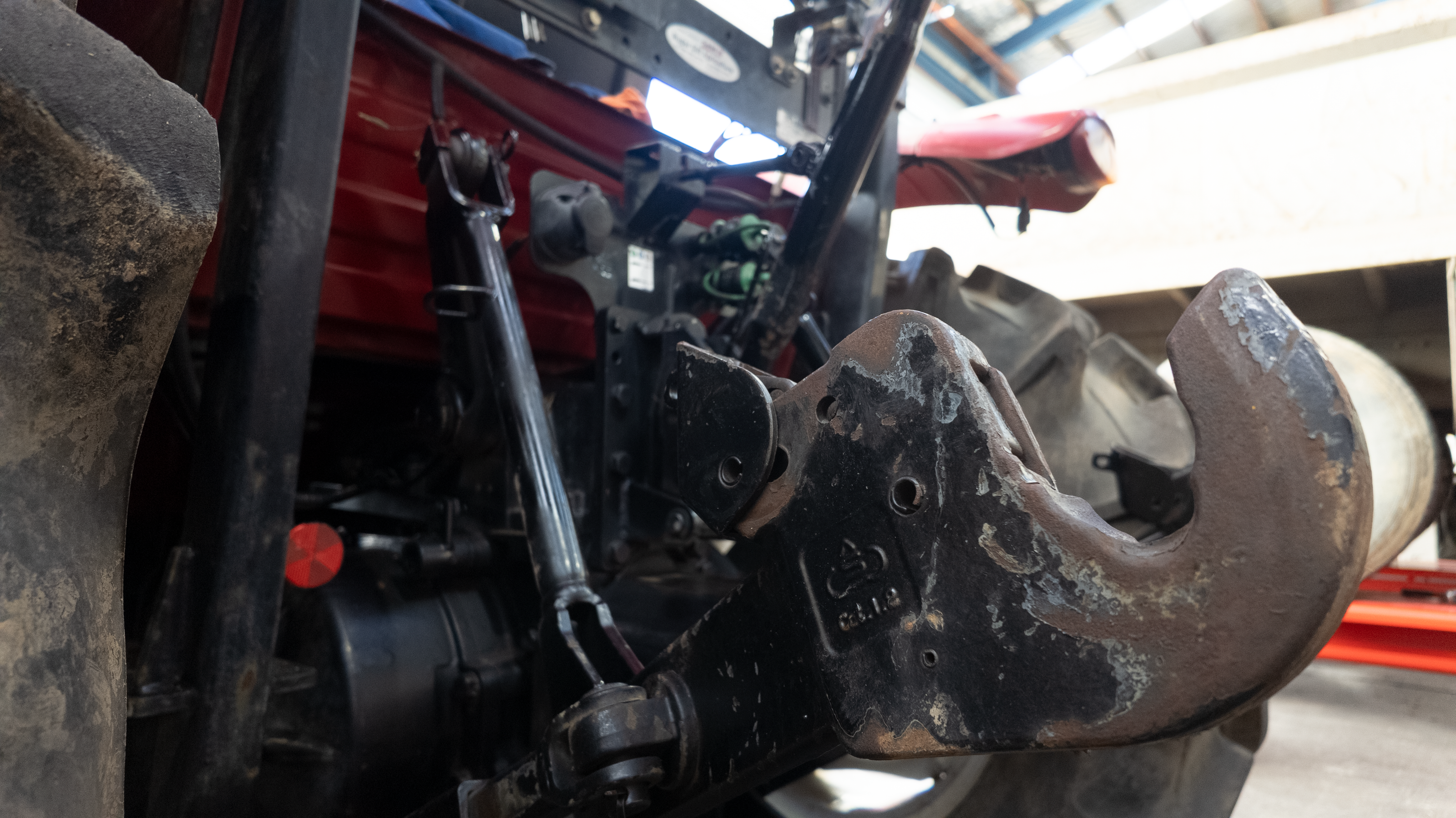 Close-up of a heavy-duty black and rusted metal tractor or farm implement hitch and linkage assembly attached to a red vehicle in a workshop setting.