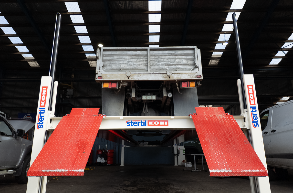 Steril Kobi truck lift hoist with red diamond-plate ramps and white support posts, shown in a diesel workshop with a vehicle elevated inside.