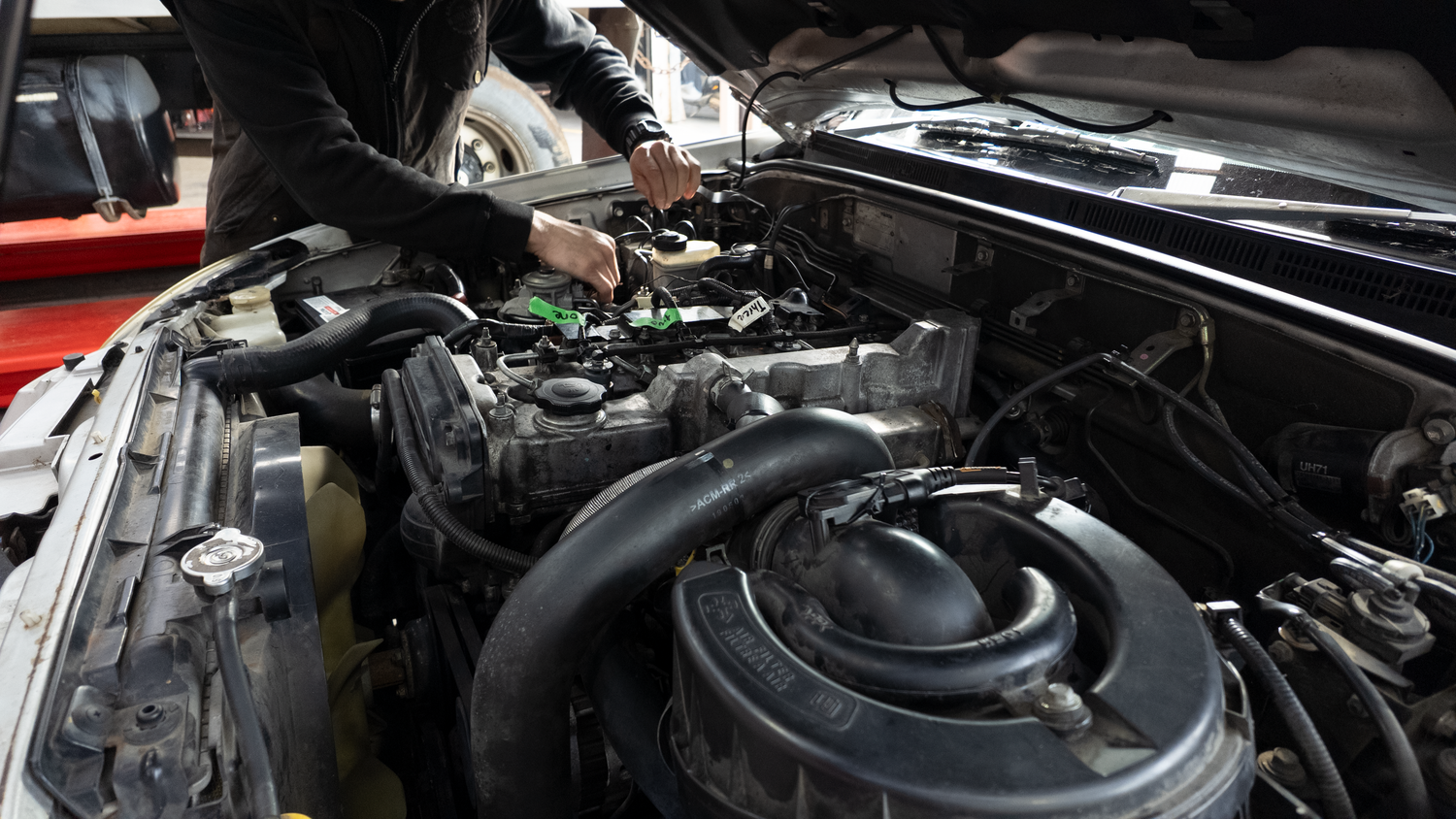 A close-up view of a car engine bay with the hood open, showing various engine components and hoses. A person in a black jacket is leaning over the engine, working on it in what appears to be a garage or workshop setting.