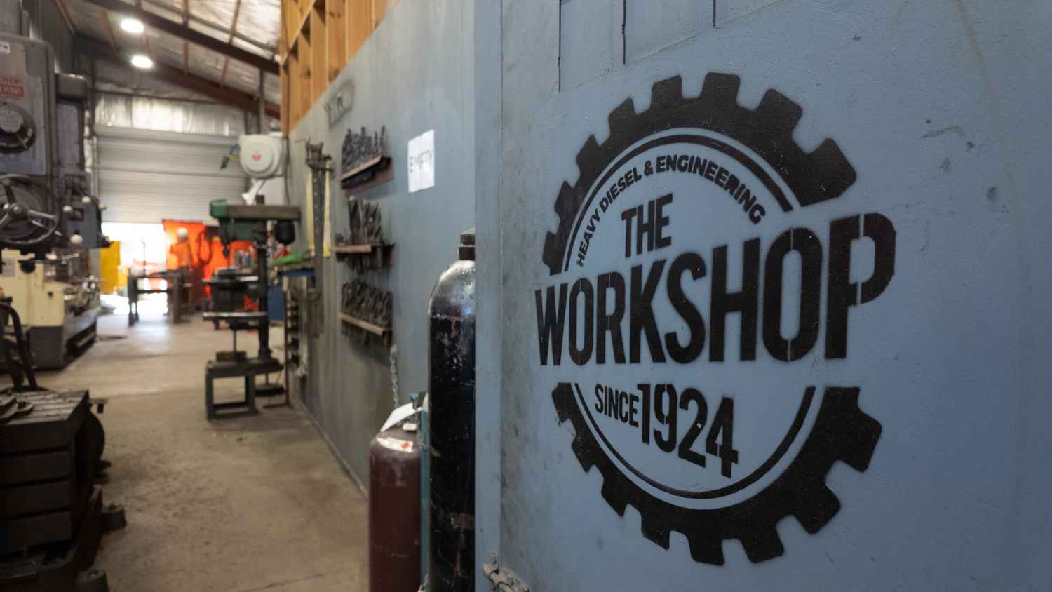 A large black metal sign reading 'THE WORKSHOP SINCE 1924' with a gear outline, mounted on a light gray wall inside an industrial workshop. Machinery, tools, and equipment are visible in the background.