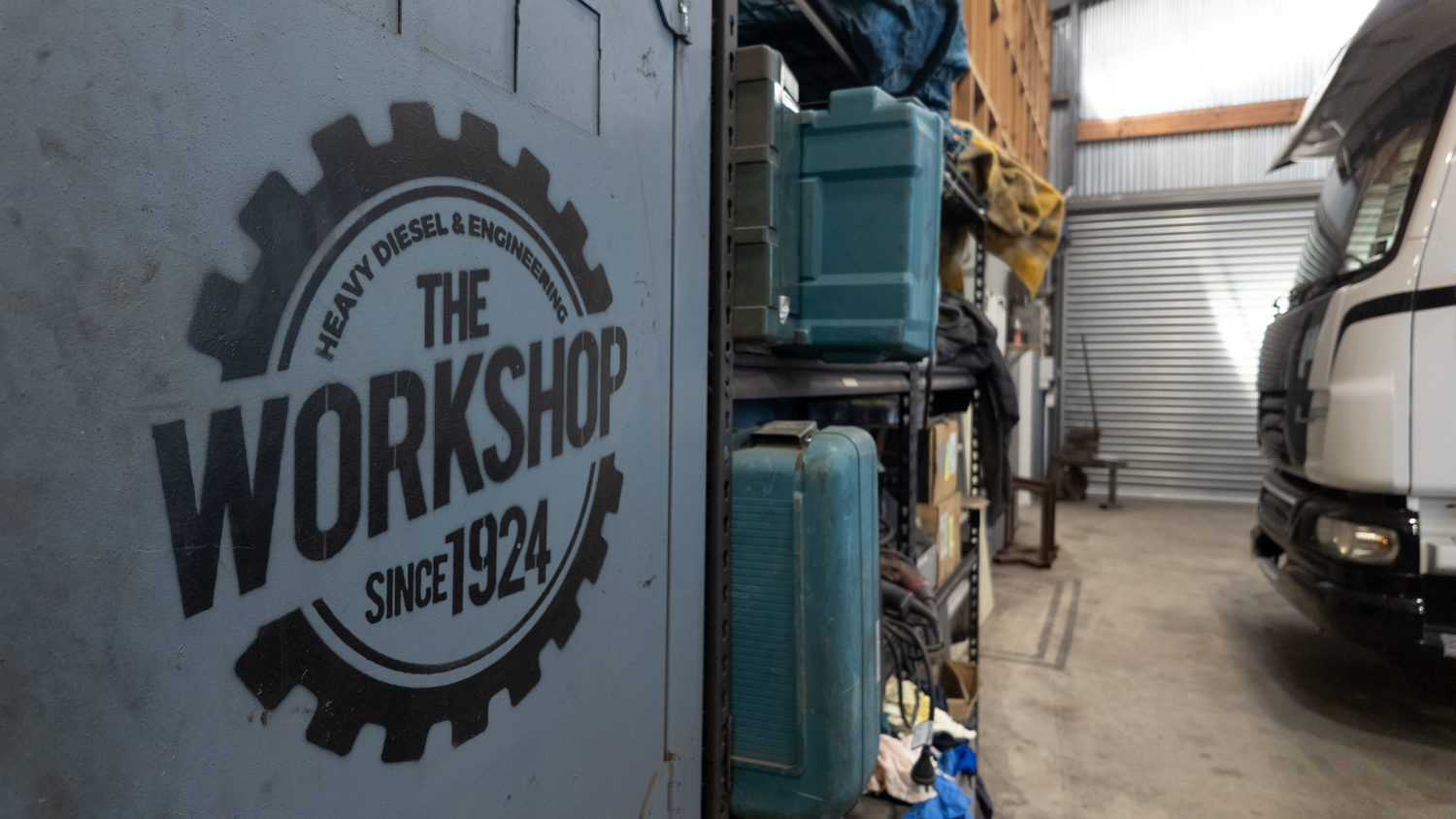 A large white industrial container or van door features a bold black circular logo reading 'THE WORKSHOP SINCE 1924' with gear graphics. The scene is set inside a workshop or garage with shelving, storage bins, and part of a truck visible in the background.